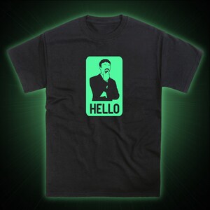 May include: Black t-shirt with a green glow-in-the-dark graphic of a man with a mustache, wearing a suit and tie, with the word "HELLO" underneath.