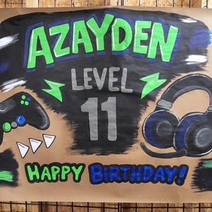 May include: A hand-painted banner with the name "AZAYDEN" in green letters outlined in blue, above the word "LEVEL" and the number "11". A black controller and headset are depicted, along with the words "HAPPY BIRTHDAY!"
