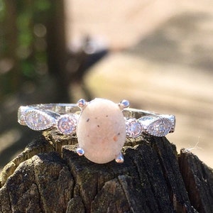 May include: A silver ring with a white oval-shaped stone set in the center. The stone is surrounded by smaller clear stones. The ring is sitting on a piece of wood.