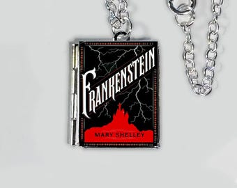 May include: A silver chain necklace with a pendant shaped like a book. The book pendant has a black cover with the title "Frankenstein" in white, lightning bolts, and a red silhouette of a castle. The author's name, Mary Shelley, is printed at the bottom.