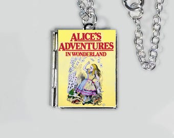 May include: A silver-colored necklace with a pendant shaped like a small book. The book cover is yellow with the title "ALICE'S ADVENTURES IN WONDERLAND" in red text. The book illustration shows Alice in a blue dress.