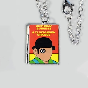 May include: A silver chain necklace with a rectangular pendant featuring the book cover of "A Clockwork Orange" by Anthony Burgess. The pendant has a red background with the book title and a stylized illustration of a man with a bowler hat.