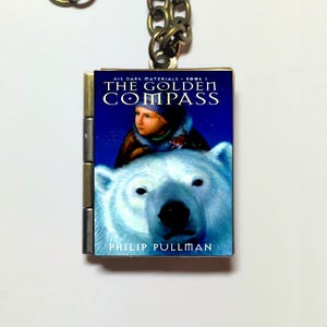 Könnte beinhalten: Eine Halskette mit einem silbernen Anhänger in Form eines Buches. Der Umschlag zeigt einen blauen Hintergrund mit weißen Sternen, den Titel "The Golden Compass" und eine Illustration eines Kindes, das auf einem Eisbären reitet.