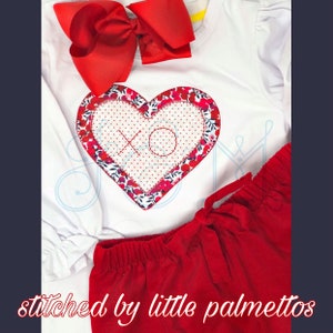Può includere: Un completo composto da camicia bianca a maniche lunghe e pantaloncini rossi. La camicia presenta un'applicazione a forma di cuore con motivi floreali e a pois, e le lettere "XO". È attaccato un grande fiocco rosso. Il testo "stitched by little palmettos" è in basso.