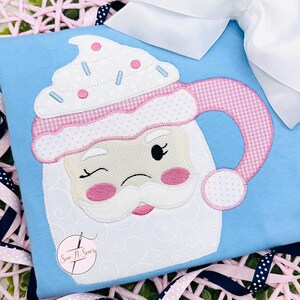 May include: A white mug with a pink and white gingham handle and a pink and white whipped cream top. The mug has a Santa face with a pink nose and a white beard. The mug is on a light blue background.