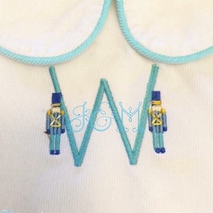 May include: A white embroidered fabric with a blue and white letter W and two blue toy soldier figures on either side of the letter.