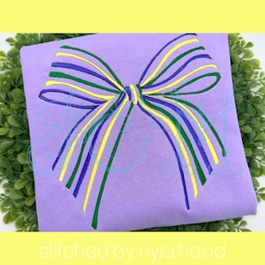 Könnte beinhalten: Ein lavendelfarbener Stoff mit einem gestickten Schleifendesign. Der Bogen hat grüne, gelbe und blaue Streifen. Der Text "stitched by nyla hood" befindet sich am unteren Rand des Bildes.