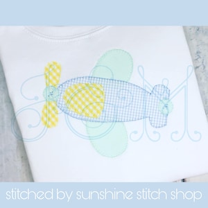 May include: A blue and yellow airplane applique with a gingham pattern. The airplane has a propeller and a window. The applique is stitched on a white shirt.