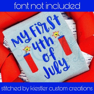 May include: A light blue t-shirt with the text "My First 4th of July" in blue embroidery. Two red and white firecracker designs are embroidered on the shirt.