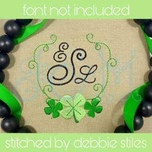 May include: A green shamrock embroidery design with the letters "S & L" in a black cursive font inside a wreath. The design is stitched on a beige fabric.