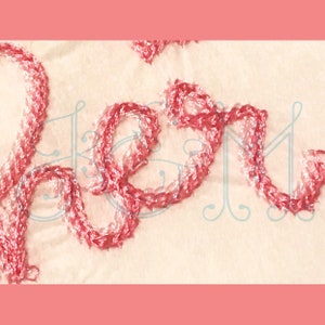 以下が含まれることがあります： 白い布地にピンクの糸で刺繍された「ママ」の文字のクローズアップ。