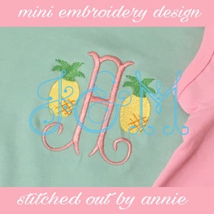Op de afbeelding: Een lichtblauw en roze raglan shirt met een aangepast borduurontwerp. Het ontwerp toont de roze letter "H" geflankeerd door twee gele ananassen met groene toppen. De tekst "mini embroidery design" en "stitched out by annie" is ook zichtbaar.