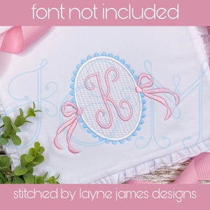 May include: A white fabric with a pink and blue embroidered monogram in a circle with a bow. The text "stitched by layne james designs" is at the bottom of the fabric.