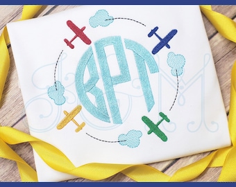 Puede incluir: Una camisa blanca bordada con un monograma circular azul rodeado de aviones y nubes. El monograma son las letras "EPT". El texto "stitched by the threaded needle" está en la parte inferior de la imagen.