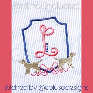 May include: An embroidered design featuring a monogrammed letter "L" in a pink script font inside a blue and white shield with two brown dogs on either side of the shield. The dogs are facing each other and have their tails curled. A pink bow with blue accents is below the shield.