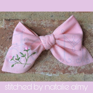 May include: A pink fabric bow with a delicate floral embroidery design. The embroidery features small pink flowers and green leaves.