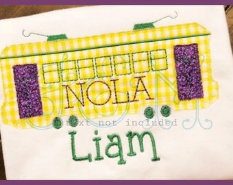May include: A yellow and white gingham embroidered design of a New Orleans streetcar with the word "NOLA" and the name "Liam" stitched below. The design is on a white t-shirt.