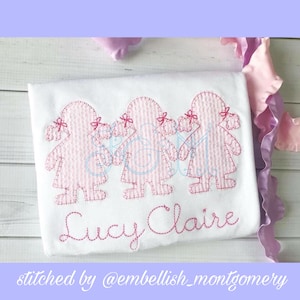 May include: A white t-shirt with three pink embroidered figures of girls holding hands. The text "Lucy Claire" is embroidered below the figures.