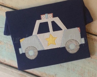 May include: Navy blue shirt with an appliqued police car design. The police car features a white body, blue plaid accents, a yellow star, and a red and white checkered light. The wheels are gray and blue. The shirt is made of soft fabric.