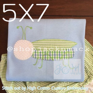 May include: A light blue baby onesie with a green and white gingham grasshopper applique. The onesie is 5x7 inches and has the text "@shopjackmack" and "J&M" embroidered on it. Stitch out by High Cotton Custom Embroidery.