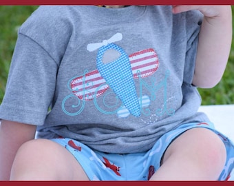 Puede incluir: Una camiseta gris con una aplicación de avión rojo, blanco y azul. El avión tiene un cuerpo azul y blanco a cuadros y alas rayadas rojas y blancas. La camiseta tiene el texto "stitched by @ashleymade1" en la parte inferior.