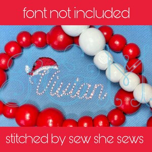 May include: A blue fabric with a red and white beaded garland and a red and white embroidered design that says "Olivia" with a Santa hat on top. The text "stitched by sew she sews" is at the bottom of the image.