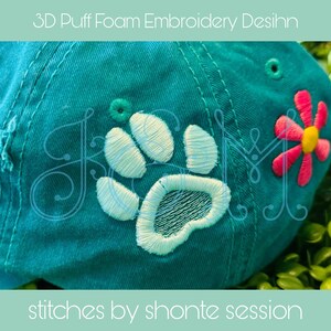 May include: A turquoise blue baseball cap with a white paw print embroidered on the front. The paw print is 3D and has a puffy texture. The cap also has a pink flower with a yellow centre embroidered on the side. The text "stitches by shonte session" is printed on the bottom of the image.