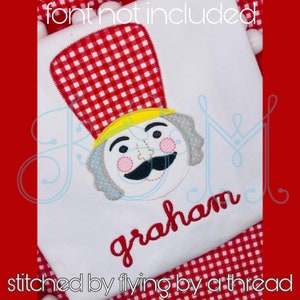 May include: A white shirt with a red and white checkered hat, a gray and yellow nutcracker face, and the name "graham" embroidered in red.