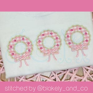 May include: Three pink and green embroidered wreaths with pink bows on a white shirt. The shirt is on a woven wicker surface. "stitched by @blakely_and_co" is written below the image.