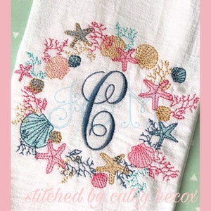 May include: Embroidered wreath with a blue letter C in the center. The wreath is made of coral, seashells, and starfish in shades of pink, blue, gold, and white. The text "stitched by cathy hecox" is at the bottom of the image.