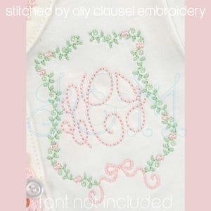 May include: Embroidered design of the word "Grace" in a pink and blue script font, surrounded by a green floral wreath with pink flowers. The design is stitched on a white fabric.