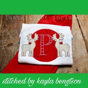 Op de afbeelding: Een witte romper met een rode cirkel en een witte letter "P" erin. Twee witte rendieren staan aan weerszijden van de cirkel. De romper ligt op een rode stof. De tekst "letters not included" staat bovenaan de afbeelding. De tekst "stitched by kayla bengtson" staat onderaan de afbeelding.