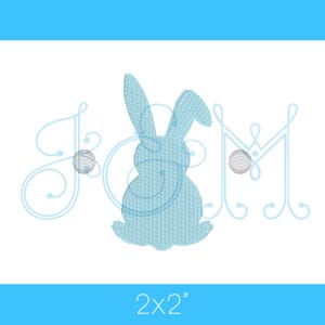 Peut inclure: Un motif de lapin brodé bleu clair avec le texte "JCM" et deux petites boules décoratives. Le motif mesure 5,08 x 5,08 cm. L'image a un fond blanc avec une bordure bleue.