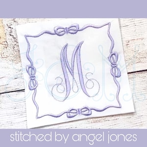 Può includere: Un quadrato di tessuto bianco con un monogramma ricamato in viola in una cornice quadrata con fiocchi ad ogni angolo. Il monogramma è la lettera "M".