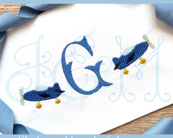 Op de afbeelding: Borduurwerk met de letter "G" in een blauw handschrift met twee blauwe vliegtuigen die eromheen vliegen. De vliegtuigen hebben gele wielen en propellers. Het ontwerp is geborduurd op een witte stoffen achtergrond.