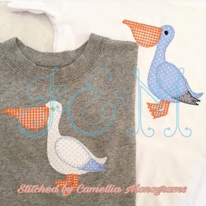 May include: Two appliqued pelican designs on grey and white fabric. The pelicans are blue and white with orange beaks and gingham accents. The text "Stitched by Camellia Monograms" is at the bottom of the image.
