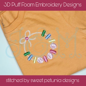 May include: Orange t-shirt with a colorful embroidered design. The design features a wreath-like shape with the word "HELLO" in the center. The text "3D Puff Foam Embroidery Designs" and "stitched by sweet petunia designs" are also visible.