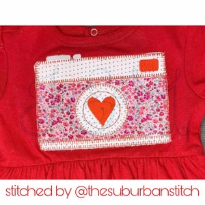 May include: Red baby dress with a decorative camera applique. The camera features a floral print, a red heart, and white polka dots. The text "stitched by @thesuburbanstitch" is at the bottom of the image.