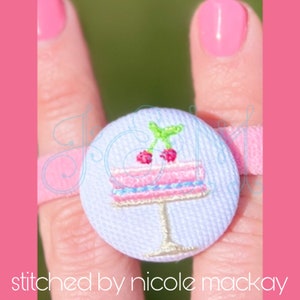 May include: A light blue fabric button with a pink and white embroidered cake with two red cherries on top. The button is attached to a pink elastic hair tie.