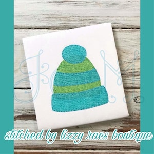 May include: A blue and green embroidered beanie with a pom pom on top. The beanie is stitched on a white fabric background.