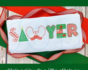 May include: A white onesie with the name "Sawyer" embroidered in colorful festive letters. The letters are decorated with Christmas-themed designs, including candy canes, a Christmas tree, mittens, and a wrapped gift. The onesie is folded and displayed on a red and white striped ribbon.