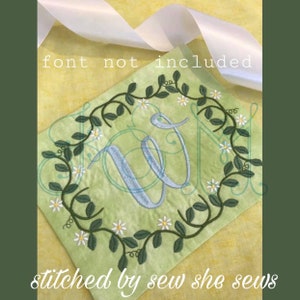 May include: Embroidered design of a letter W surrounded by a wreath of green leaves and white flowers. The text "stitched by sew she sews" is at the bottom of the image.