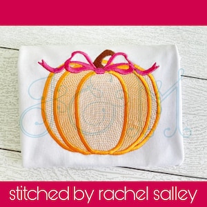May include: Embroidered pumpkin with a pink bow on a white t-shirt. The pumpkin is orange with a light orange center and a brown stem.