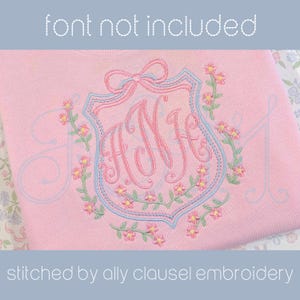 May include: Pink sweatshirt with a floral monogram design. The design features a shield shape with a bow at the top, a floral wreath, and the letters "ANH" in a cursive font. The text "font not included" and "stitched by ally clausel embroidery" are also visible.