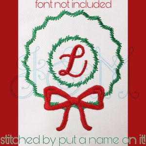Puede incluir: Una corona verde con un lazo rojo y la letra L en el centro. La corona está bordada sobre un fondo de tela blanco.