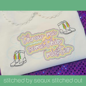 Op de afbeelding: Wit shirt met een Peter Pan kraag, geborduurd met de tekst "Throw me something mister" in paars, groen en geel. Twee paar laarzen met regenboogkwasten zijn ook op het shirt geborduurd.