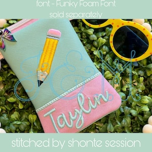 Könnte beinhalten: Ein Stiftetui aus tealfarbenem und rosafarbenem Stoff mit dem Namen "Taylor", der in einer kursivschriftlichen Schriftart gestickt ist. Auf der Vorderseite des Etuis ist ein gelber Bleistift mit einem rosa Radiergummi gestickt.