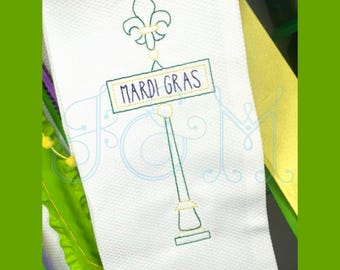 May include: Embroidered design of a green Mardi Gras sign with a fleur-de-lis on top. The sign reads "Mardi Gras" in purple thread.