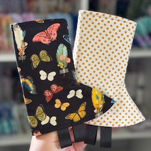 Peut inclure: Deux gants de cuisine en tissu. L'un présente un fond noir avec un motif coloré de papillons et d'éléments botaniques. Le second possède un fond blanc avec un motif répétitif de petits pois de couleur jaune moutarde.