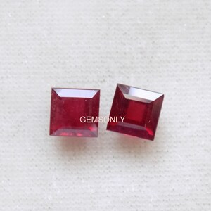 Puede incluir: Dos gemas cuadradas de color rojo intenso. Las gemas están sobre un fondo blanco texturizado. El texto "GemsOnly" es visible en las gemas. Las gemas son probablemente rubíes, con una superficie pulida y facetada.